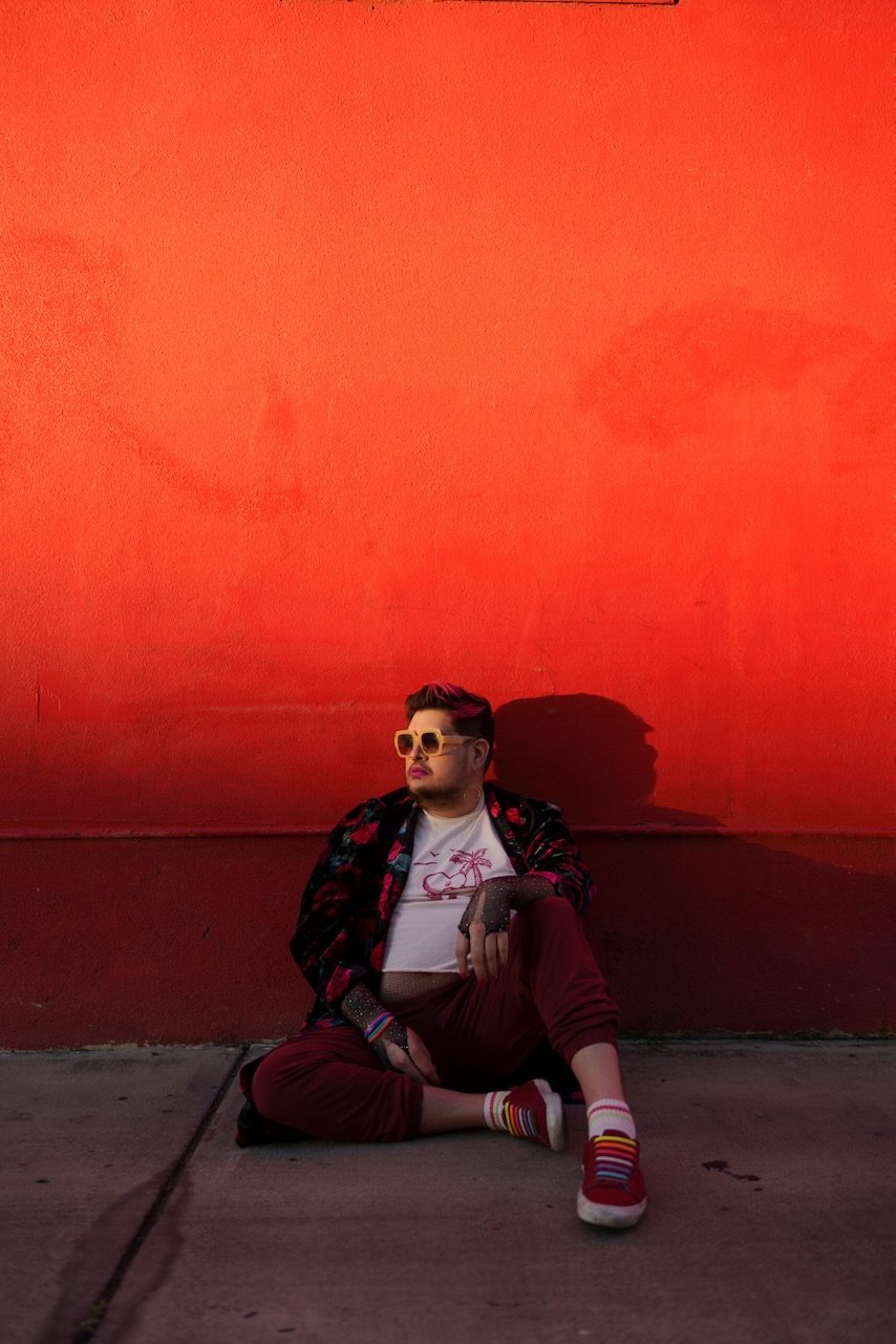Violet Paniko seated on the ground against a saturated orange-red wall, in sunglasses and a floral jacket