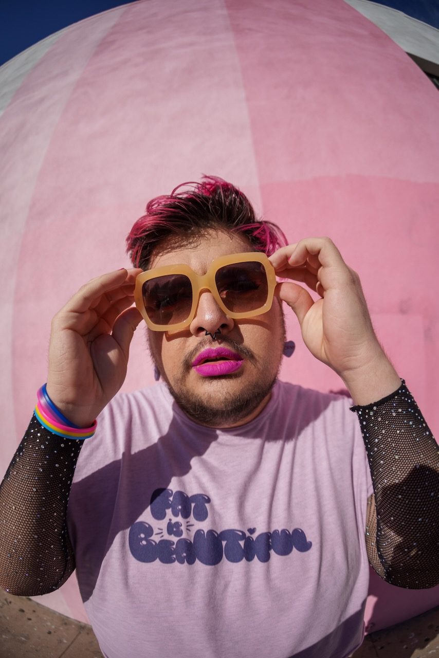 Violet Paniko in tan square sunglasses and hot pink lipstick, hands raised to the frames, against a pink dome